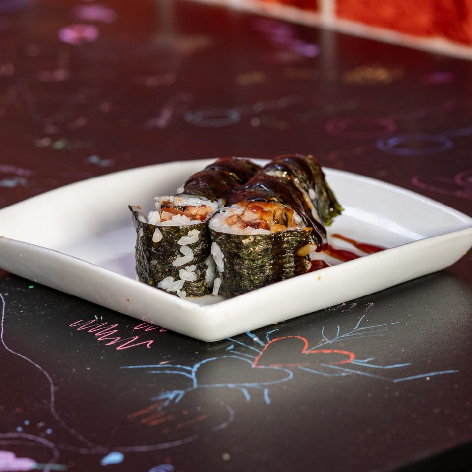 Sushi rolls with dark sauce on a white rectangular plate, on a dark table with colorful drawings.