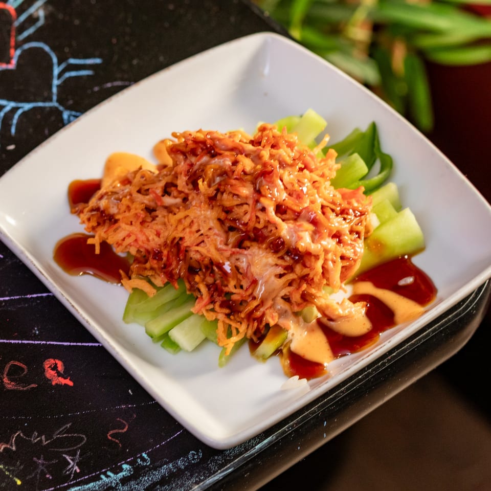 Sushi dish with shredded topping, green vegetables, and sauces on a white square plate.