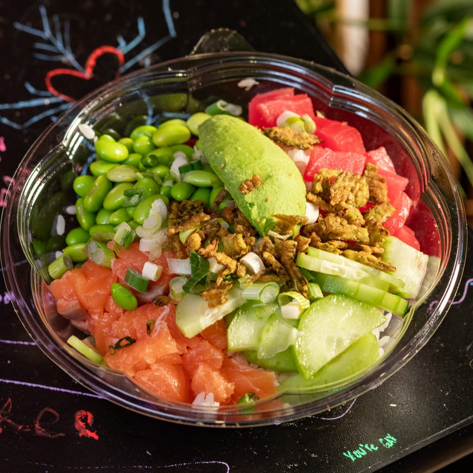 Colorful sushi poke bowl with salmon, edamame, avocado, cucumber, and watermelon on a dark surface.